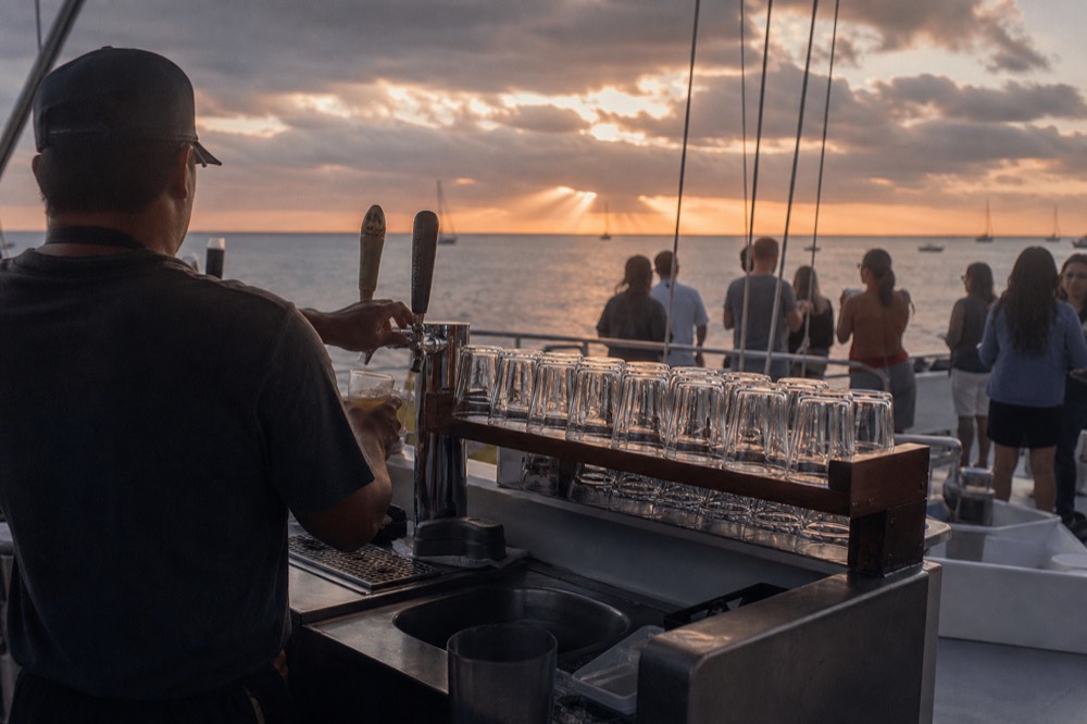 A small boat bar with beer taps and a rack of glasses, a crew member pouring while guests wait with plates in the background at dusk