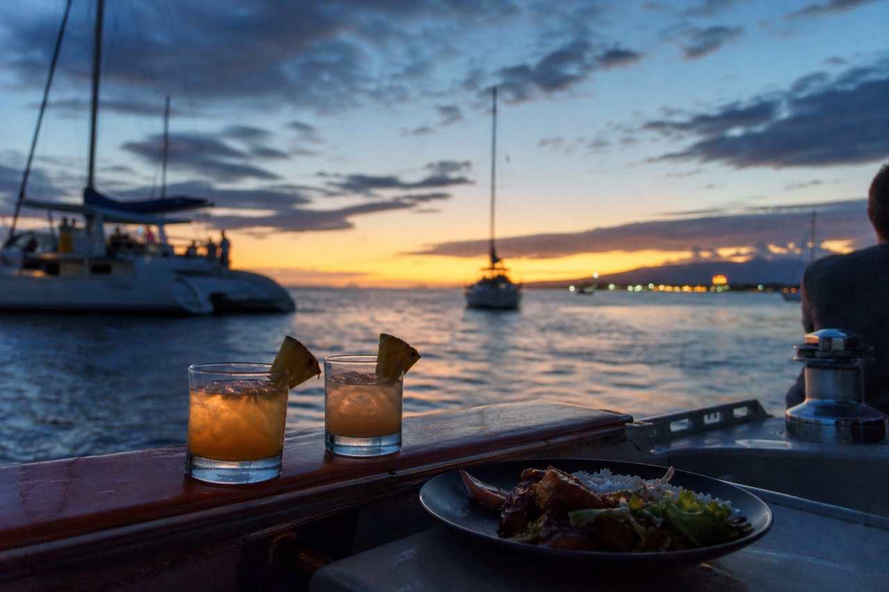 Two mai tais garnished with pineapple resting on a boat rail beside a plated dinner, low golden light across the water behind