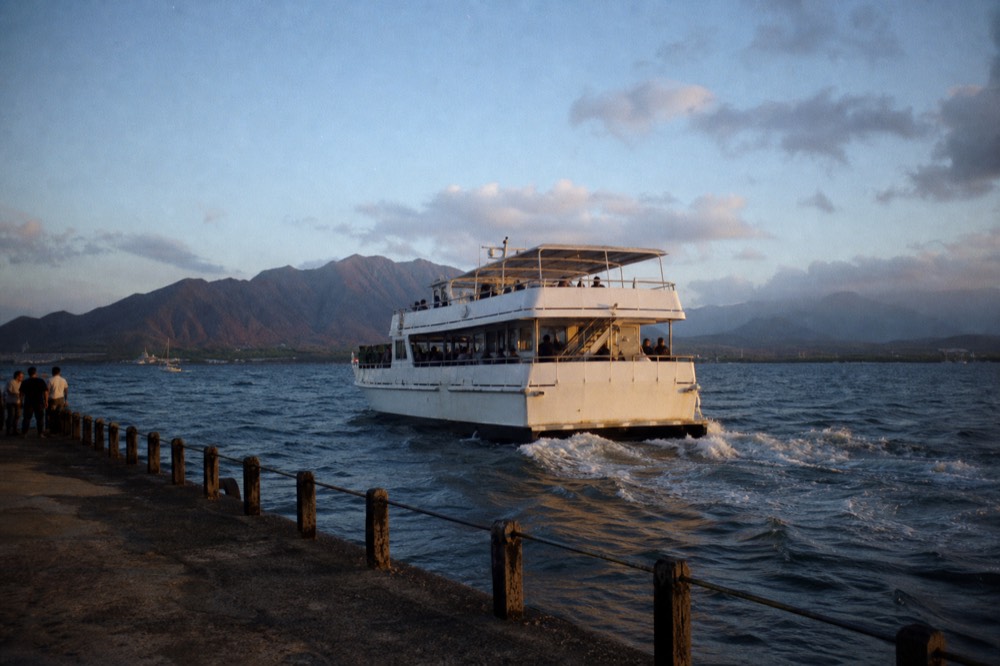A double-deck cruise boat leaving a small harbor into a windy afternoon chop, whitecaps on the bay, mountains hazy behind