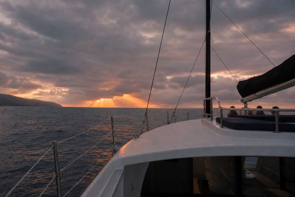 Wide upper deck of a multi-level catamaran at dusk, empty rail space and covered seating below, colour breaking through cloud on the horizon