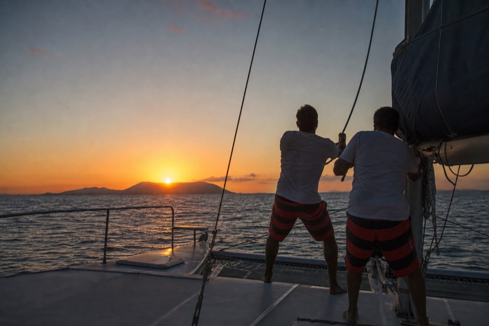 Two crew members in matching shorts hauling a mainsail halyard on a catamaran deck as the sun sits just above a distant island