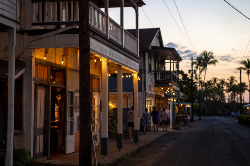 Two-storey wooden plantation-era storefronts along a narrow street at dusk, string lights over the sidewalk, a few people walking past open doorways
