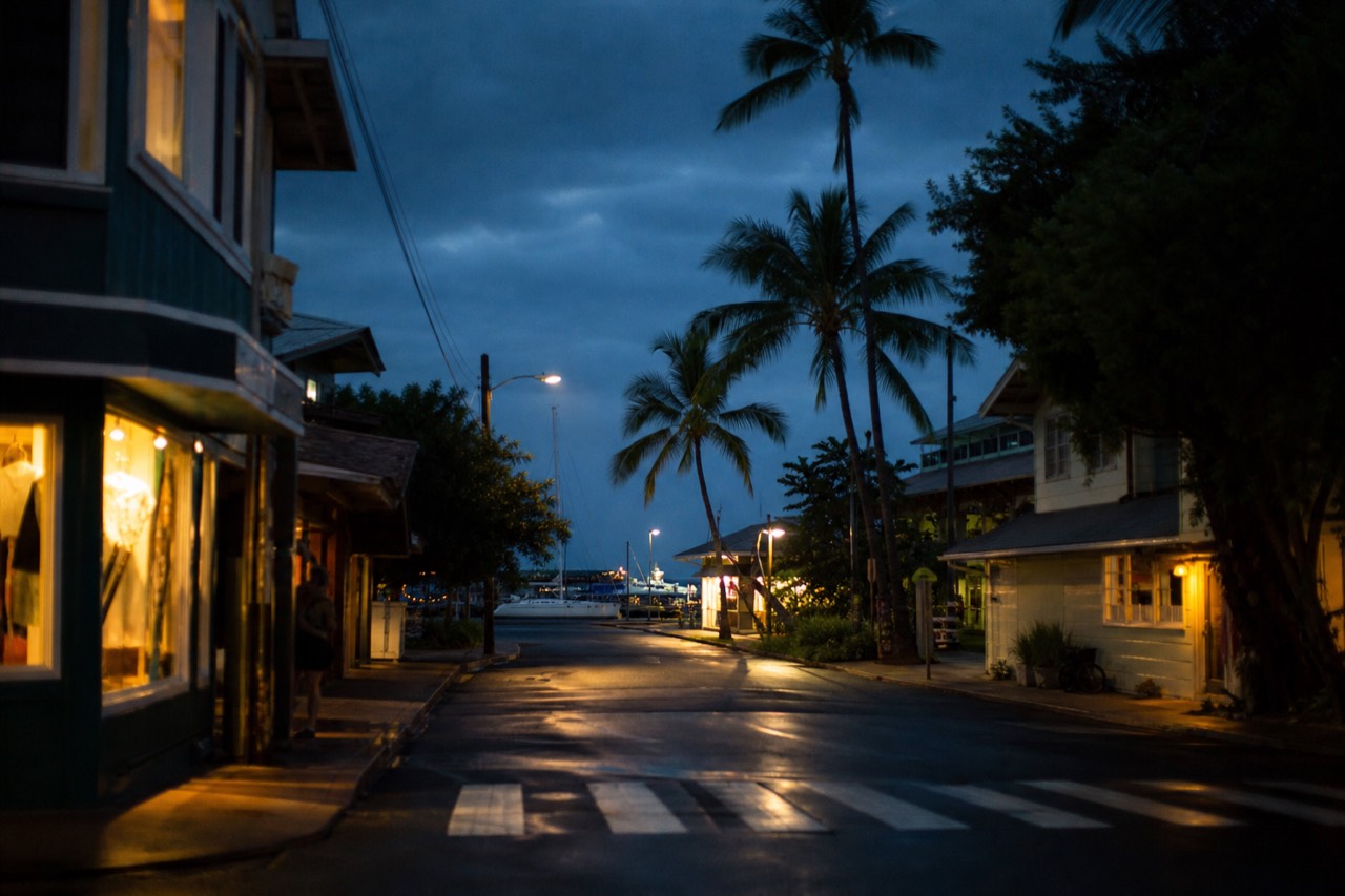A small Hawaiian town street after dark, warm light spilling from a few open storefronts onto damp pavement, palm shadows and an empty crosswalk