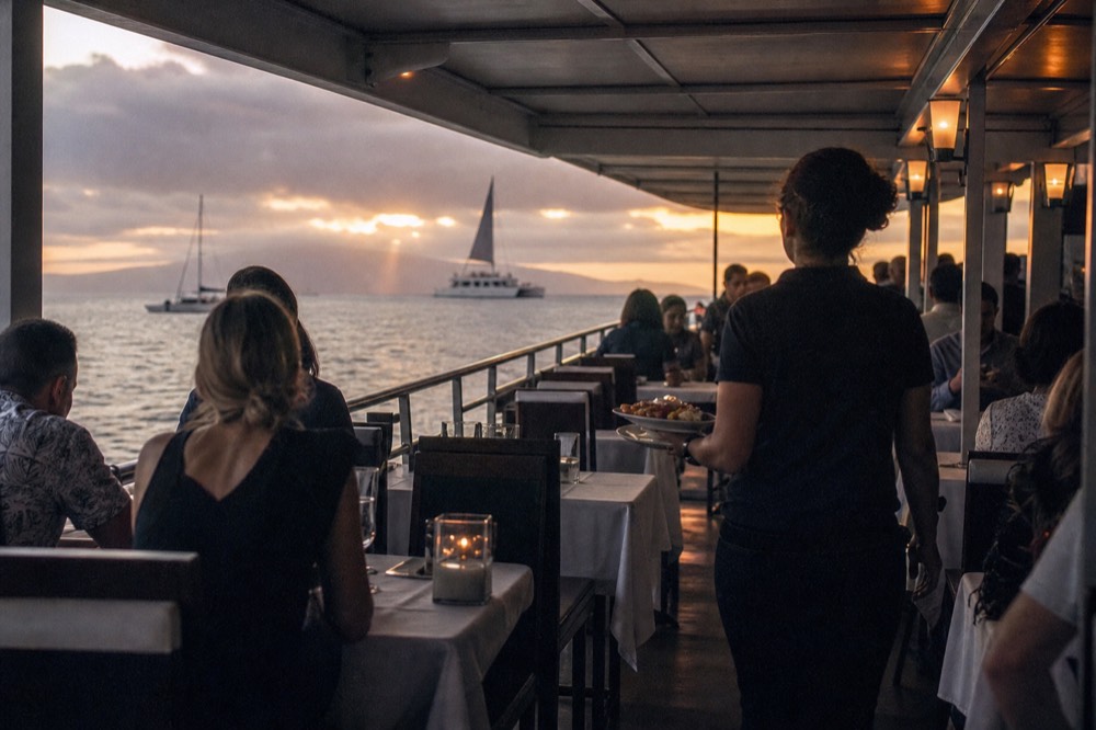 Guests seated at set tables on the covered deck of a dinner boat as a server carries plates down the aisle, evening light through open sides