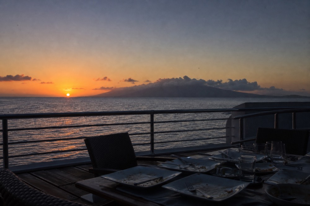 The last of the sun dropping toward a low island silhouette seen from a boat deck, empty dinner plates cleared on a table in the foreground