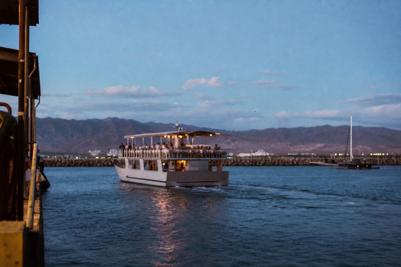 A double-deck dinner boat easing away from a harbor slip in late afternoon light, tables set on the upper deck, mountains behind the breakwater