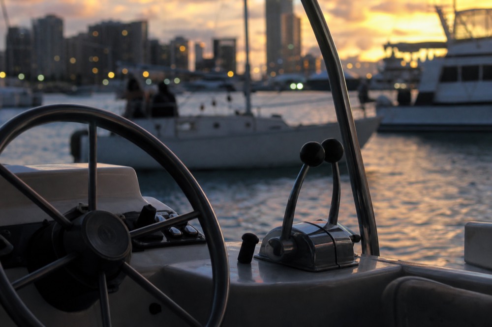 A boat wheel and throttle in close view at dusk, harbour lights out of focus behind, water reflecting the last colour