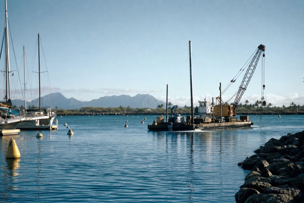 A dredging barge working inside a small boat harbour on a clear morning, crane arm over the water, buoys marking the channel
