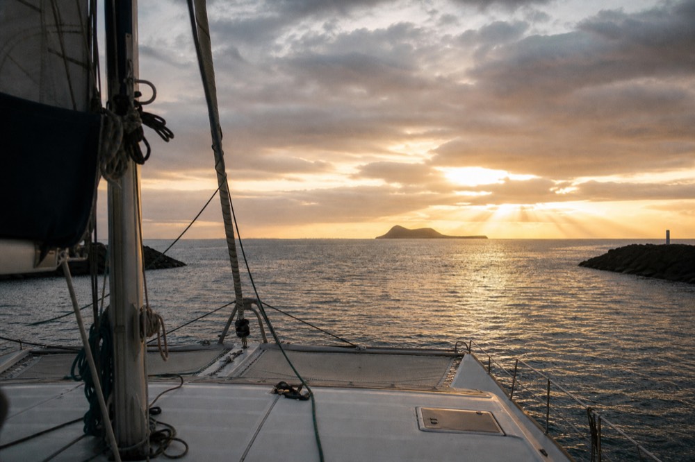 The bow of a sailing catamaran clearing a harbour entrance in late afternoon light, open channel and a distant island ahead