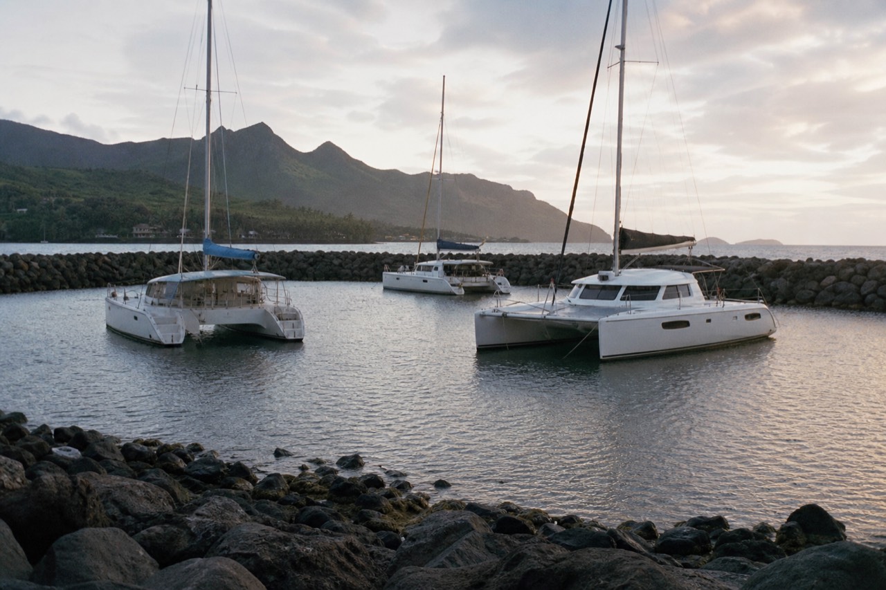 Small catamarans moored inside a harbour breakwater at dusk, calm water, a green mountain ridge rising behind the shoreline