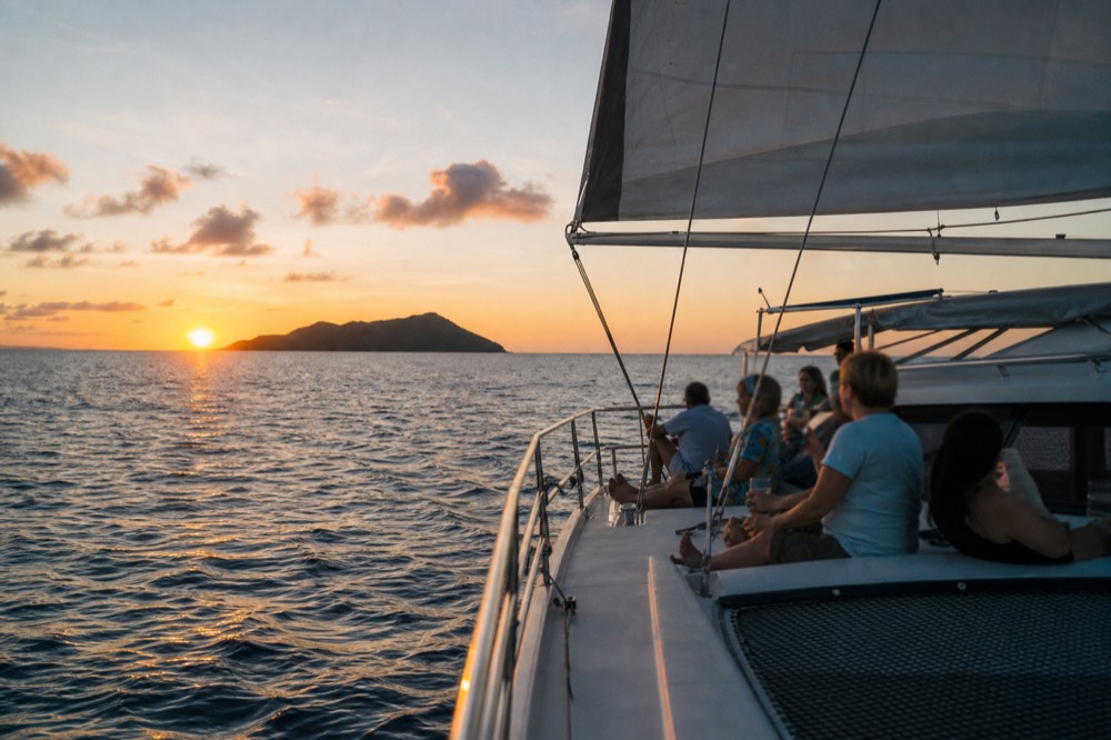 A sailing catamaran heeled slightly on calm water at sunset, the sun touching the sea beside a low dark island profile, guests along the rail with drinks