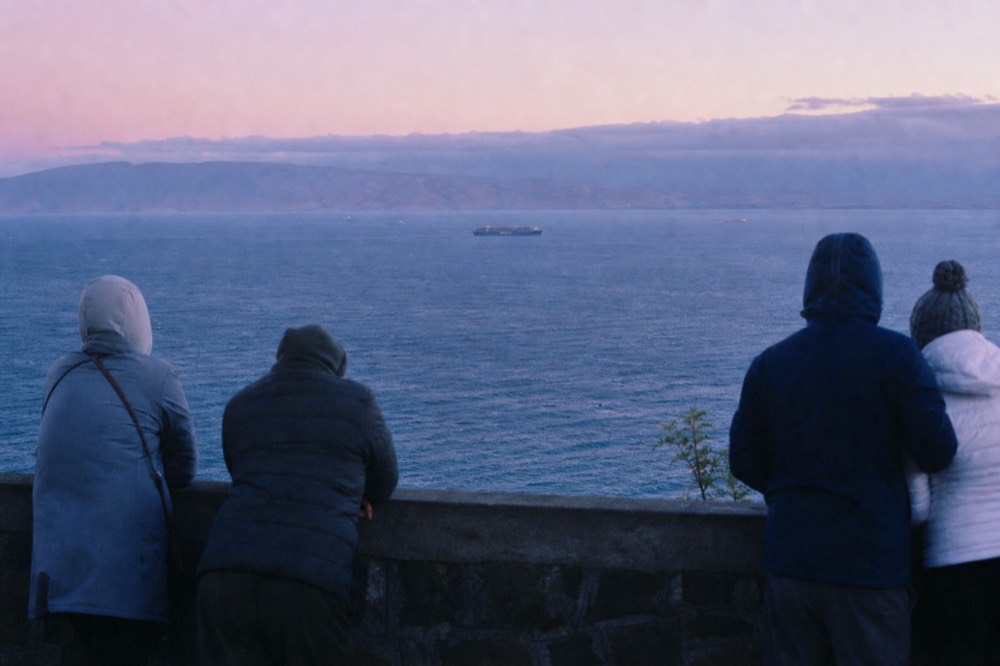 Visitors seen from behind in heavy jackets and hats at a high stone overlook rail, wind visible in their clothing, cold blue light after the sun has gone