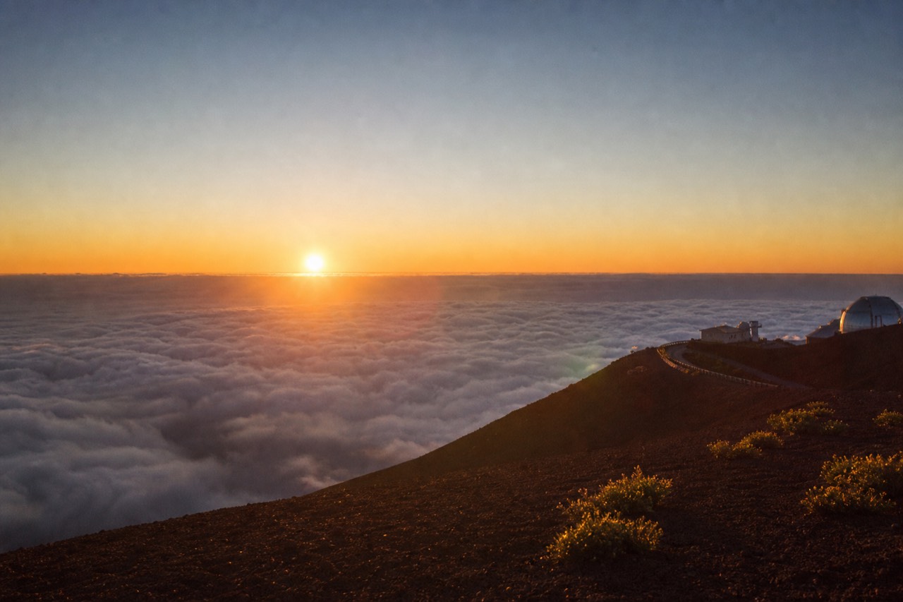 A volcanic summit ridge above a flat white sea of cloud at sunset, red cinder ground in the foreground, the sun sinking into the cloud tops rather than water