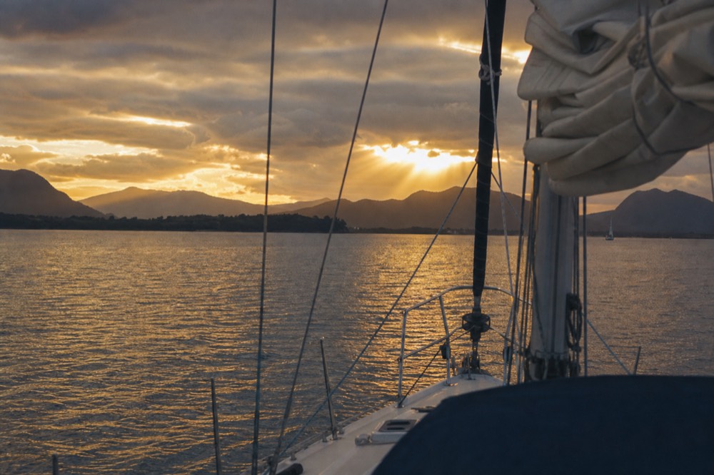 Layered island ridgelines silhouetted across a wide channel at sunset, the water pale gold, seen from the bow of a sailing boat
