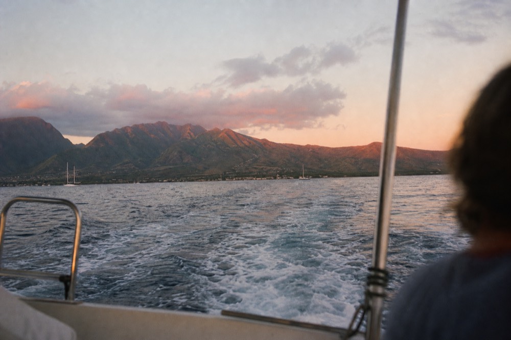 Mountain ridges above a coastline glowing pink after sunset, seen from the stern of a boat with its wake trailing toward shore