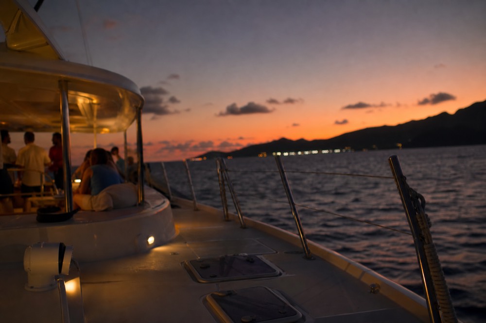 The moment just after the sun has dropped, pink and orange sky above a dark island silhouette, deck lights glowing on a returning catamaran