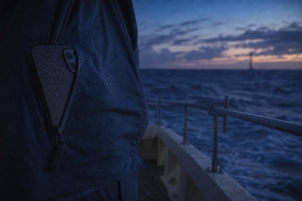 A phone tucked into the zipped pocket of a windbreaker on a moving deck, fine spray beading on the fabric under a dusk sky