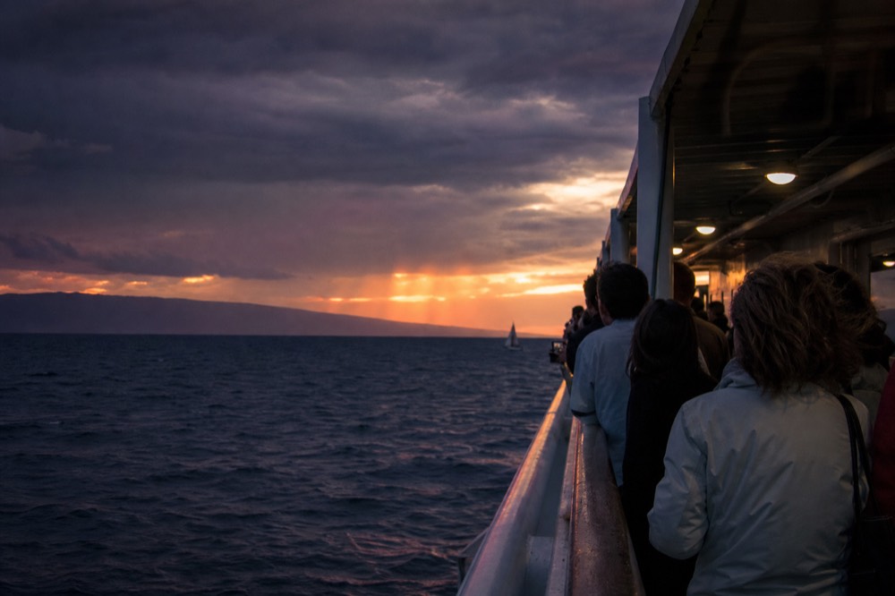 Guests in light jackets along a boat rail after sundown seen from behind, deck lights on, dark water and a purple horizon