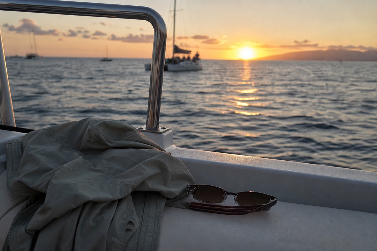 A folded light jacket and sunglasses resting on a boat bench beside a rail, low orange sun over open water beyond