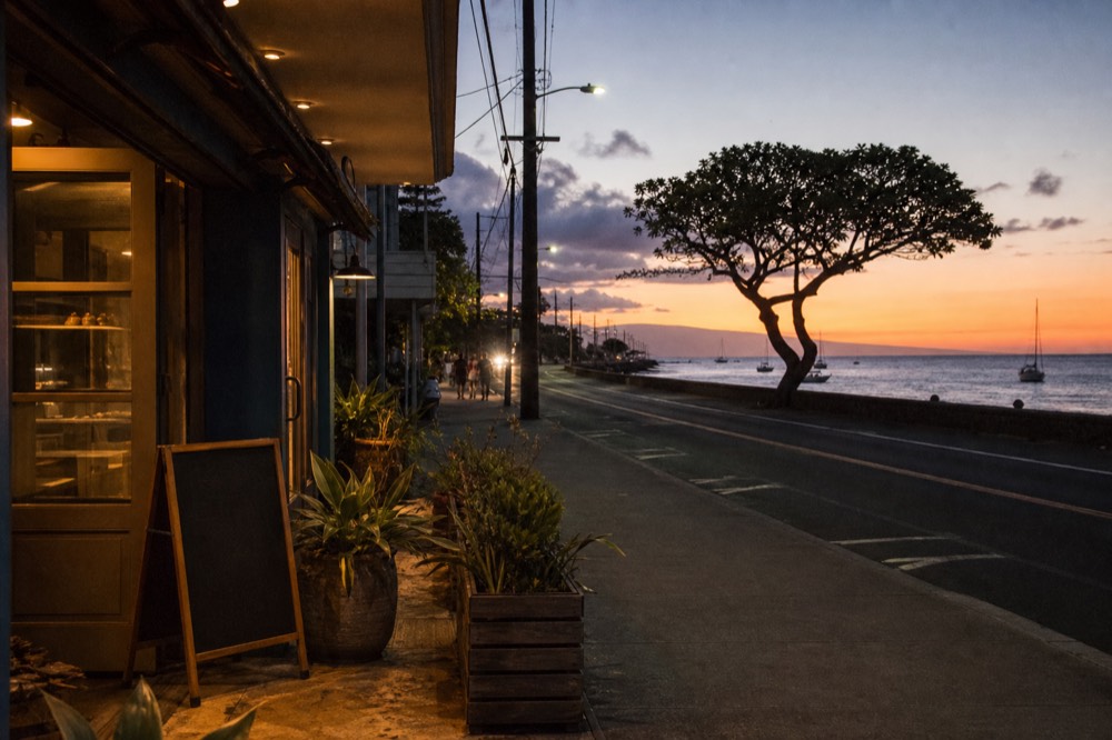 A reopened storefront on a West Maui street at early evening, warm interior light, a chalkboard menu without readable text, planters along the sidewalk