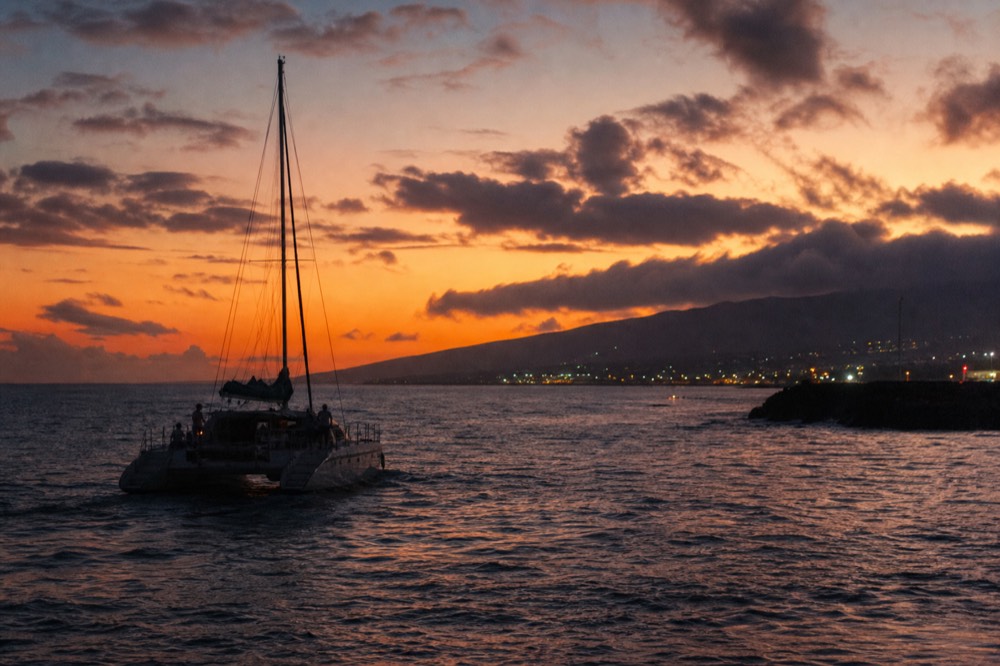 Last light over a West Maui harbor breakwater, a returning catamaran silhouetted against orange water, harbor lights beginning to show onshore