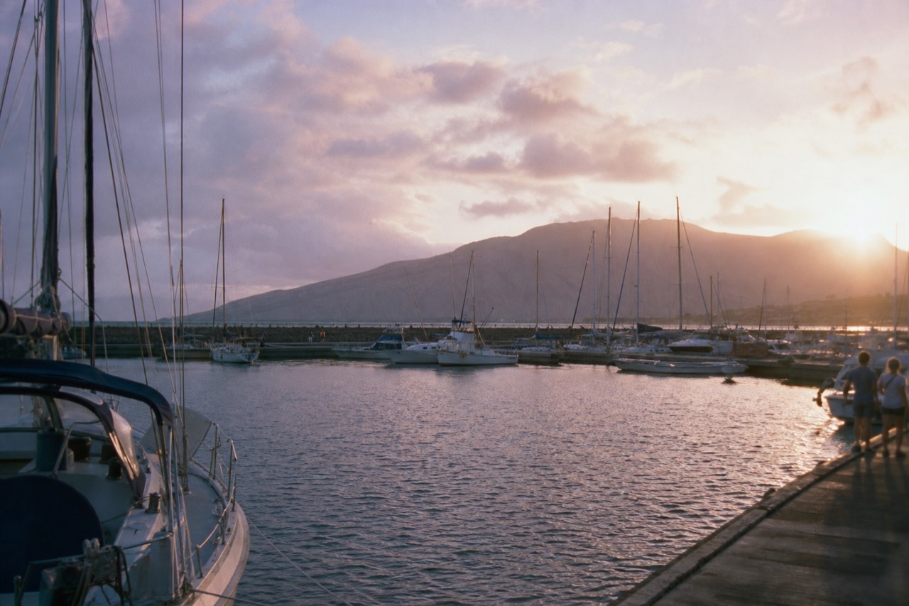 A small harbor in West Maui at dusk, masts and rigging against a violet sky, calm water inside the breakwater and a mountain ridge behind