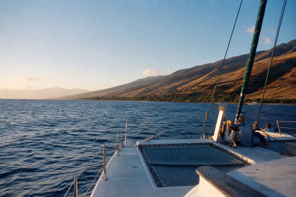 Wide view along a leeward Hawaiian coastline in late afternoon, dry golden hillsides above the shore, clear sky over open ocean to the west