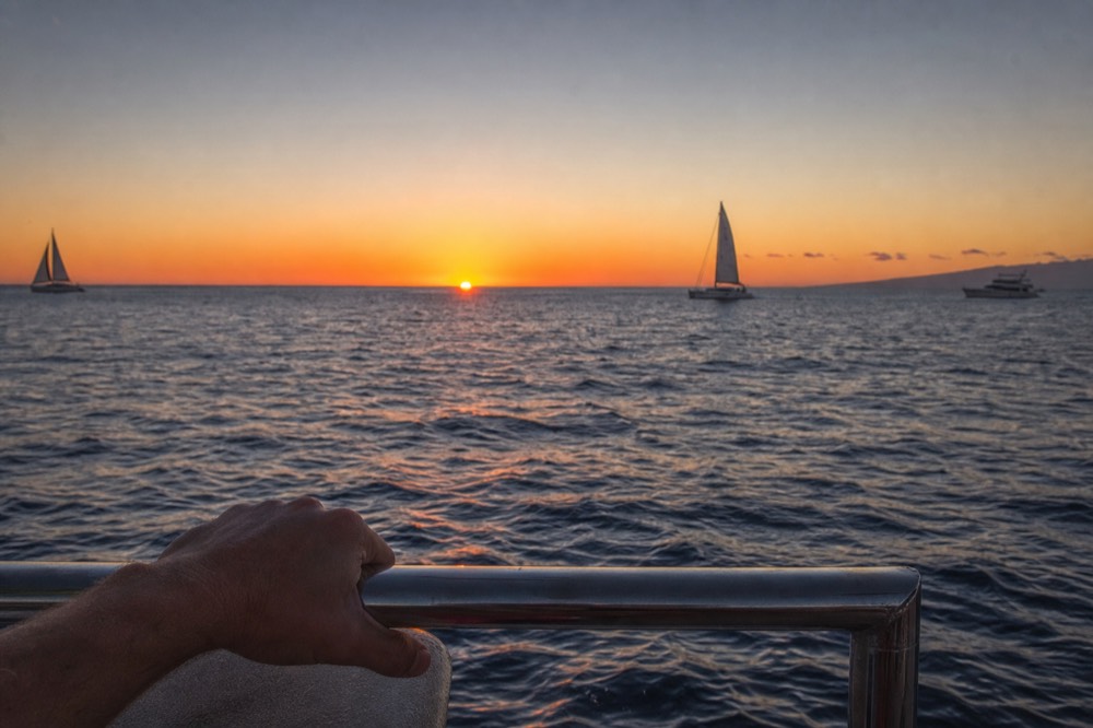 A hand resting on a stern rail with a steady flat horizon line beyond, dusk light spreading across the water