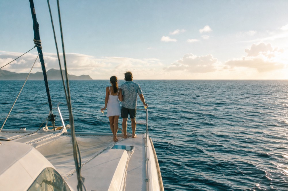 A couple standing alone at the bow rail of a catamaran holding drinks, warm backlight and empty sea filling the rest of the frame
