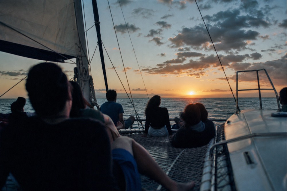 Passengers seated along the trampoline netting between the hulls of a catamaran as the sun drops toward a flat orange sea