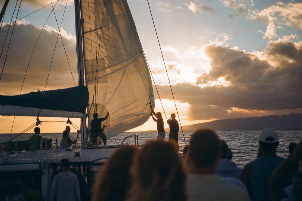 Crew hoisting a mainsail on a mid-sized catamaran, low sun backlighting the sailcloth and a scatter of guests watching from the deck