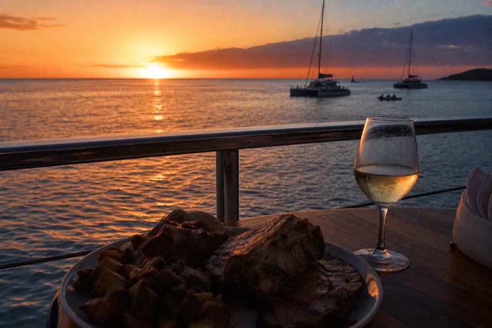 A carved prime rib plate and a glass of white wine on a small table beside an open boat rail, warm evening light on the water beyond