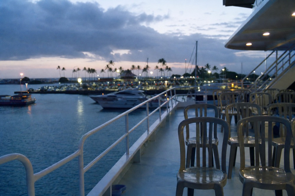 An empty upper deck of a large tour vessel at dusk with rows of plastic chairs and a bare railing, harbor lights beginning to show