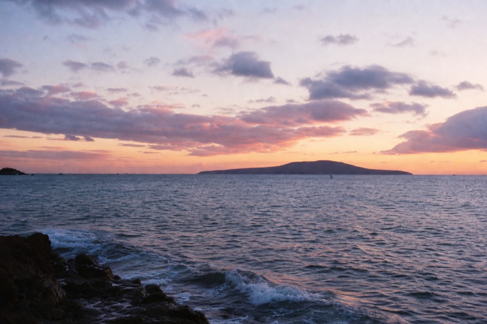 Wide open bay at sunset with a small crescent island on the horizon and layered clouds catching pink and gold light
