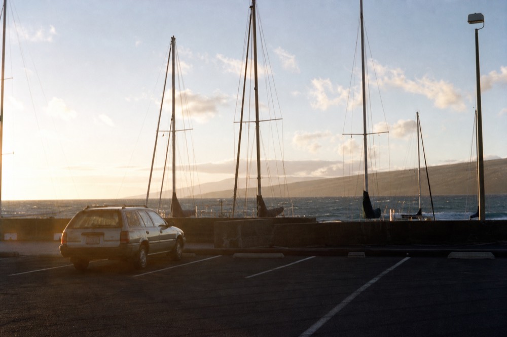 Empty harbor parking area at dusk with a single car and the masts of moored boats visible over a low wall
