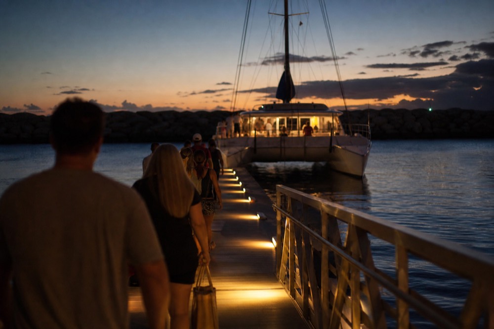 Passengers walking along a floating dock toward a lit catamaran as the last color fades from the sky behind the breakwater