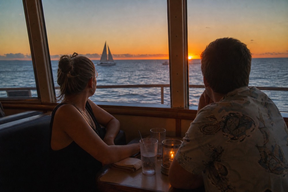 Couple seated at a window table on a double-deck boat with candle light and the sun sitting on the horizon outside