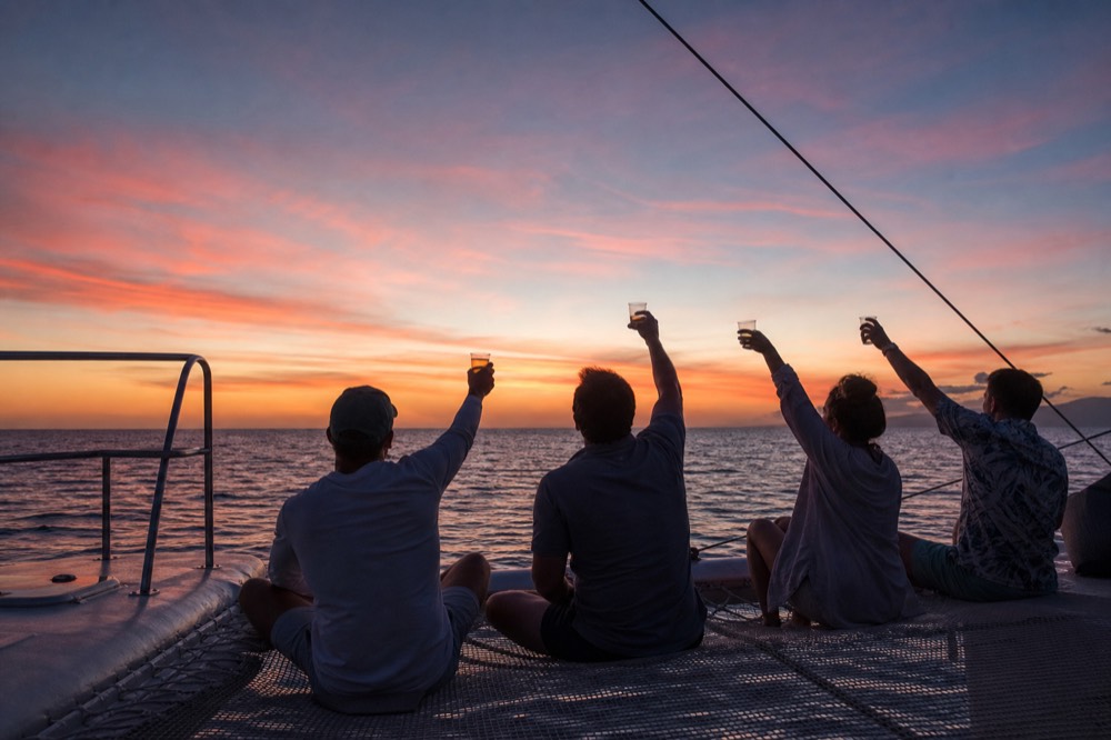 Group of friends barefoot on a catamaran trampoline with drinks raised toward a wide orange and pink sky