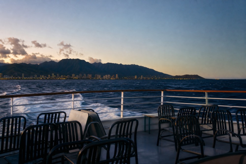 Empty deck chairs and a folded table on a returning boat with harbor lights beginning to show against a deep blue sky