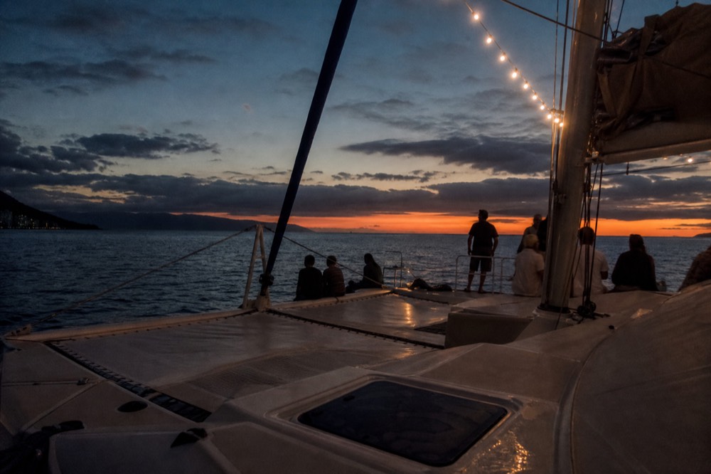 Wide deck of a returning catamaran at blue hour with string lights on and the last orange line low on the horizon