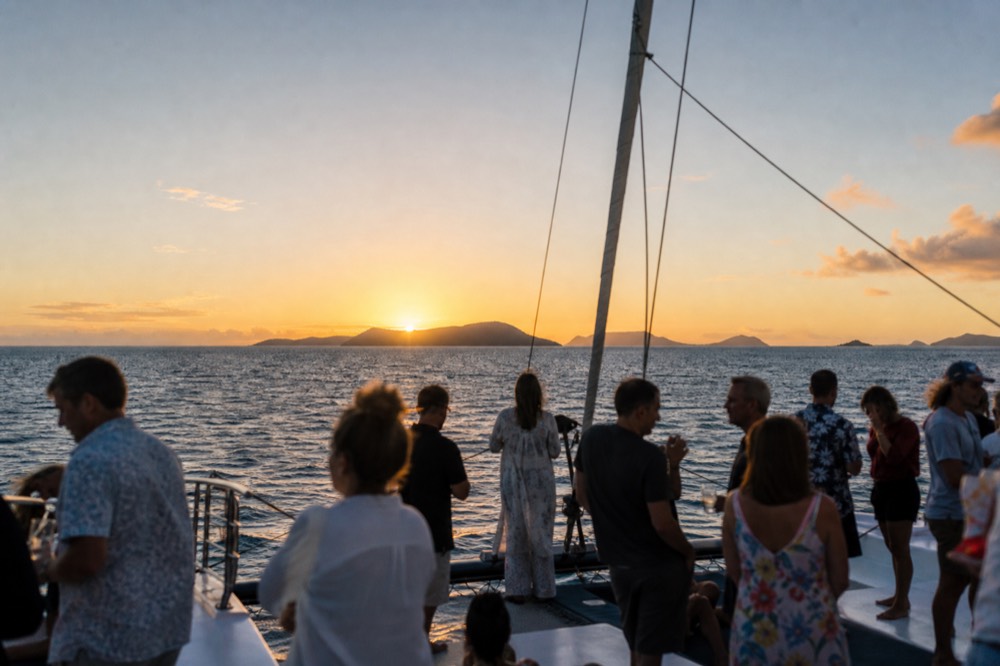 Small open-bar catamaran at golden hour with a dozen adults standing with drinks and no children aboard