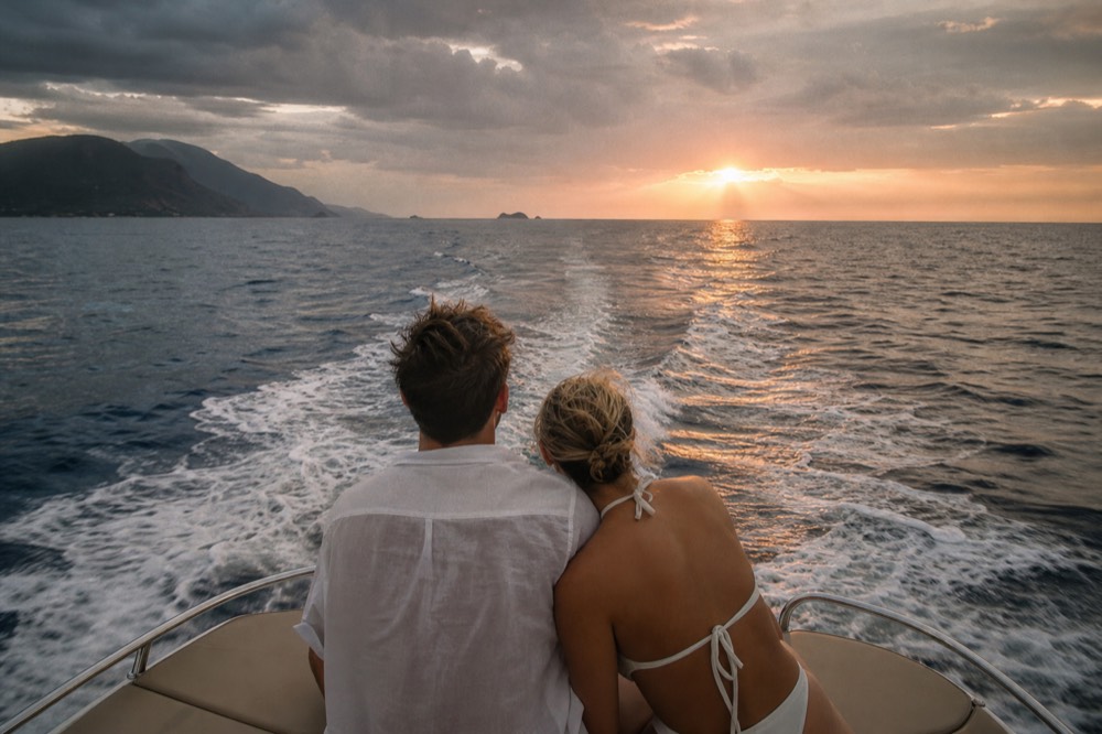 Couple alone at the bow of a small powerboat with the wake spreading behind them and low sun ahead
