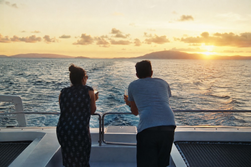 Two people leaning on the stern rail of a catamaran with drinks, wake trailing behind into a low gold horizon