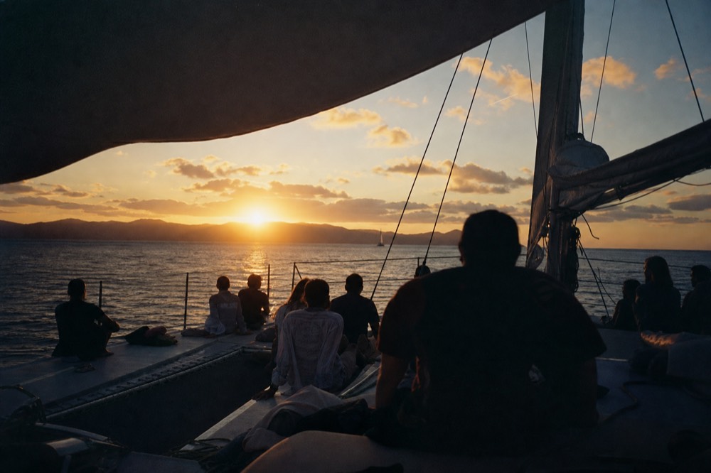 Wide view of a sailing catamaran's shadowed deck full of seated guests with the sun sitting just above a distant island ridge