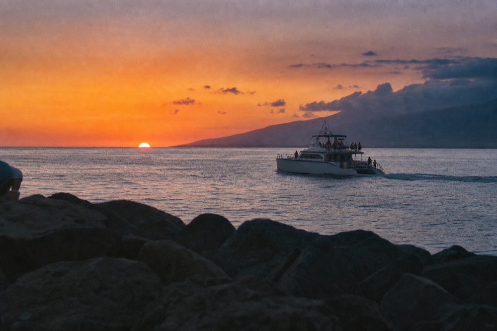 A modest twin-hull cruise boat leaving a harbor mouth in late afternoon, breakwater rocks in the foreground and mountains behind