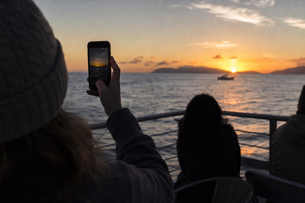 Phone screen raised toward a low sun over water with the deck rail and dark silhouettes of other passengers around it