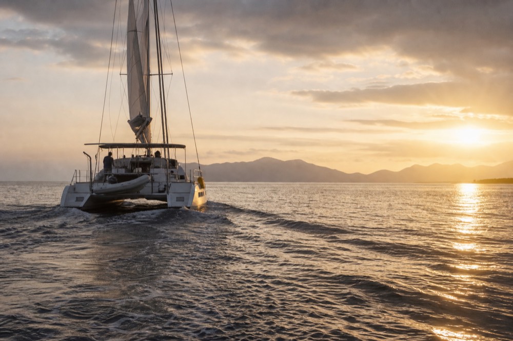 Sailing catamaran seen from astern crossing a calm channel toward a distant island ridge with the sun low on the right