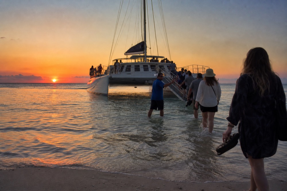 Crew member in shorts guiding barefoot passengers through shallow surf toward a catamaran bow ramp on a beach in late afternoon light