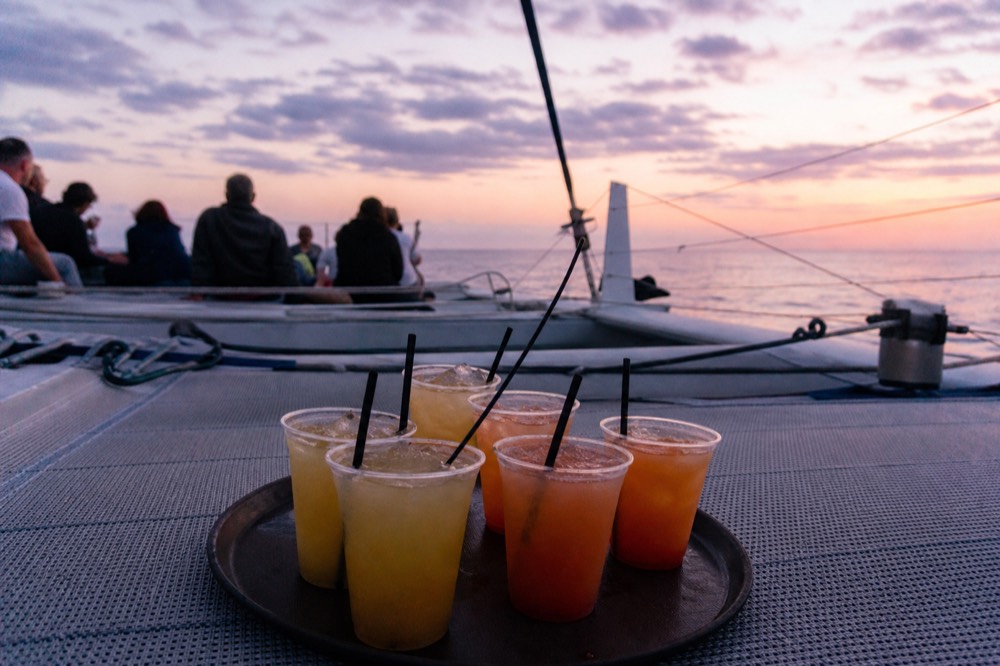 Tray of cocktails in plastic tumblers resting on a catamaran trampoline deck with rigging and orange sky behind