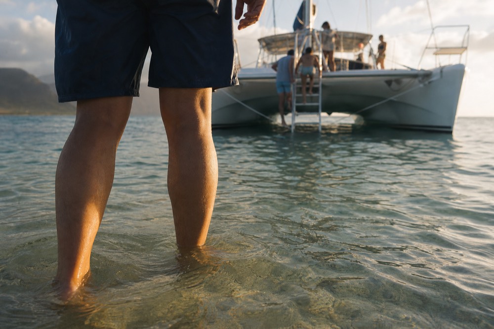 Shot from knee height in clear shallow water showing bare legs wading toward a catamaran bow ladder with soft golden light